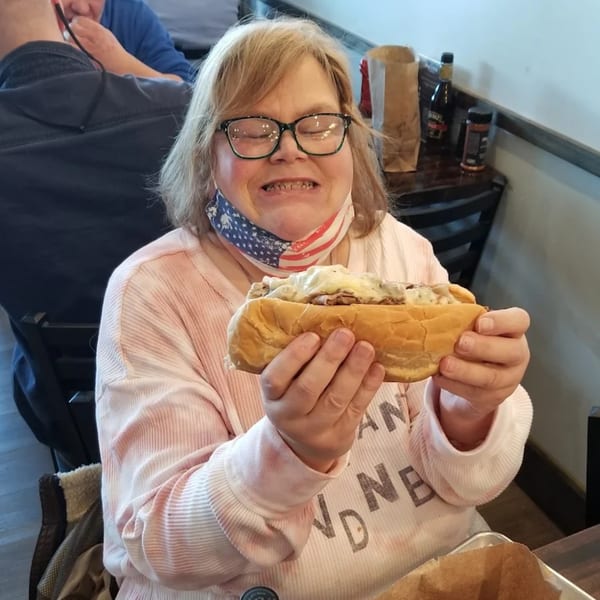 A woman with glasses and an American flag mask holds a large pit beef sandwich.