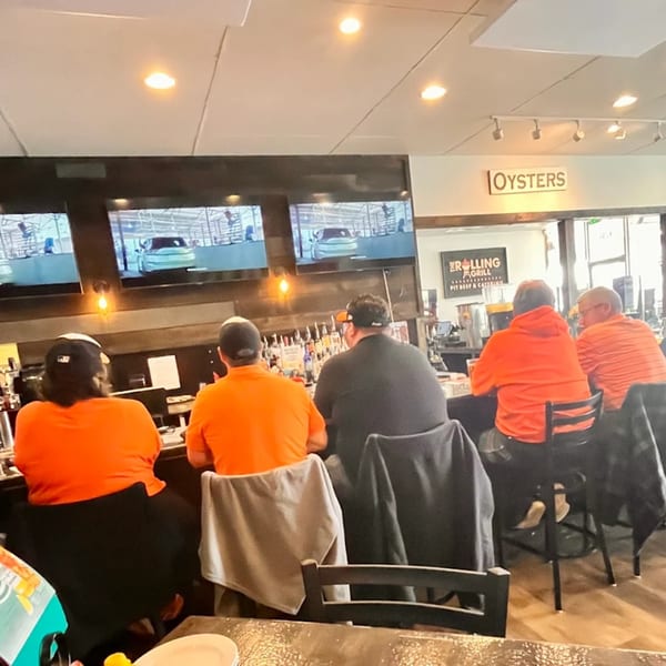 Patrons in orange shirts sit at The Rolling Grill Pit Beef bar watching TVs, with an "Oysters" sign visible.