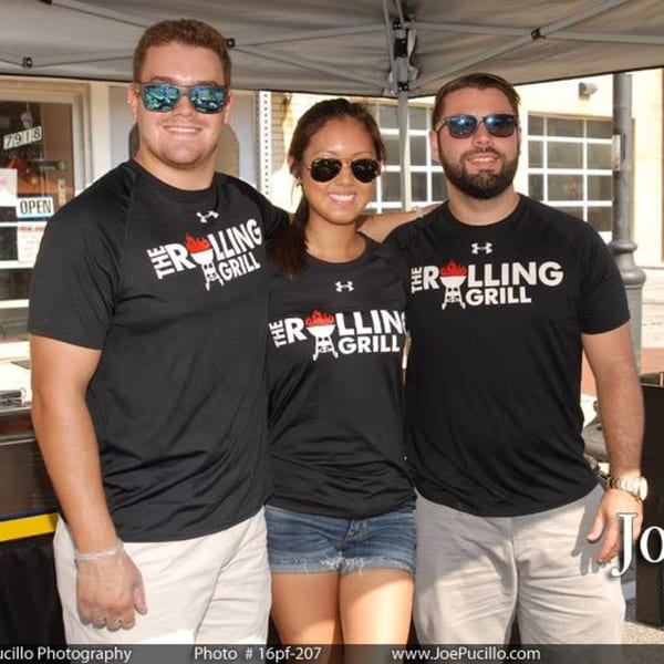 Three smiling people wearing black "The Rolling Grill" t-shirts and sunglasses stand together.
