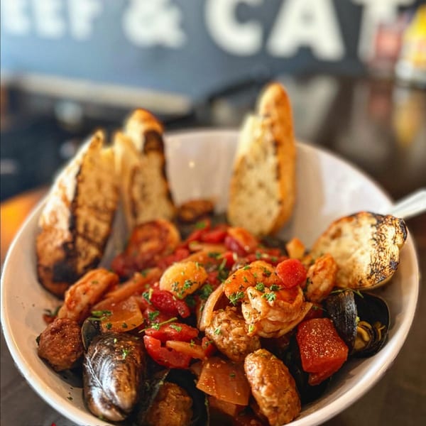 A white bowl filled with mussels, shrimp, sausage, tomatoes, and onions, served with grilled bread.