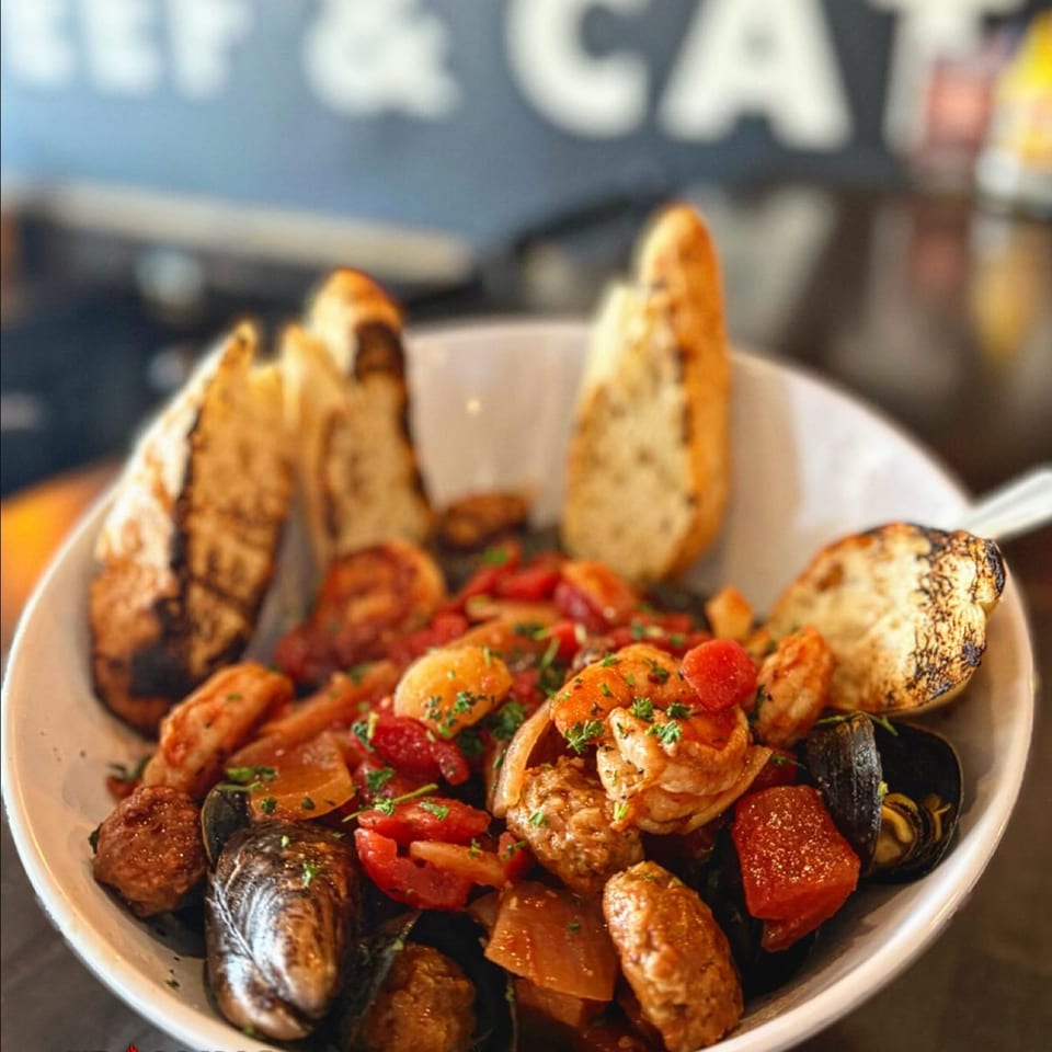 A white bowl filled with mussels, shrimp, sausage, tomatoes, and onions, served with grilled bread.