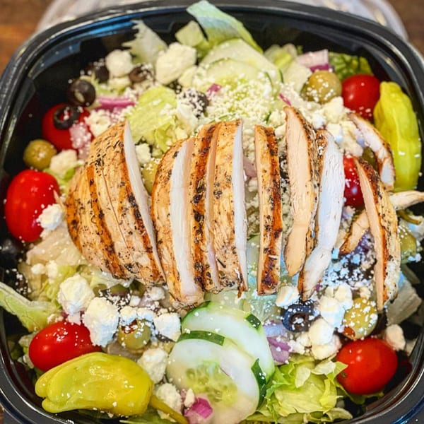 Grilled chicken salad with lettuce, tomatoes, cucumbers, olives, and feta cheese in a black bowl.