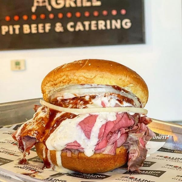 A pit beef sandwich from The Rolling Grill, topped with onions, white sauce, and BBQ sauce.