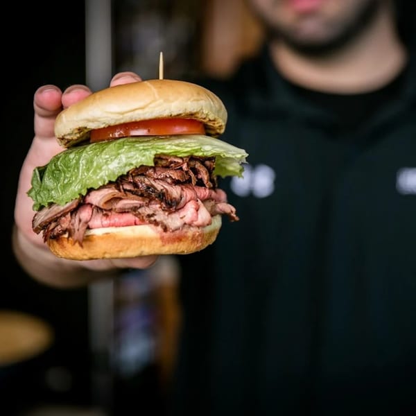 A hand holds up a delicious pit beef sandwich from The Rolling Grill, with lettuce and tomato.